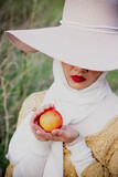 A Muslim woman in a hat sits on the field and eats peaches. Red lips. beauty