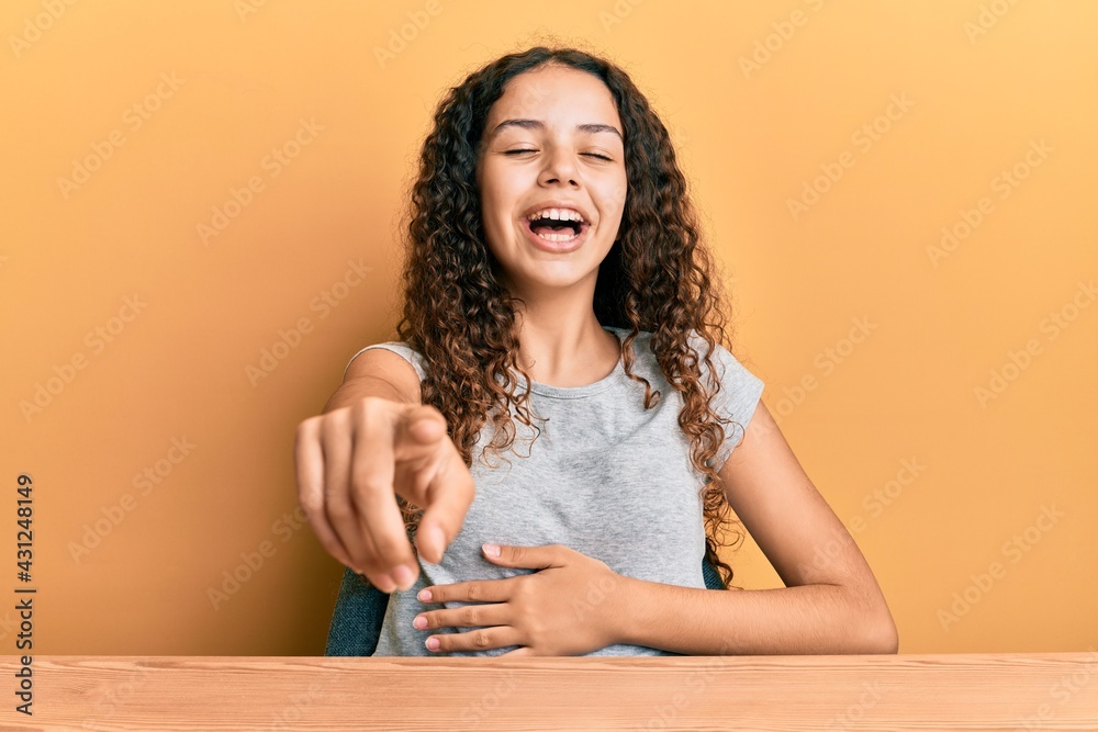 Teenager hispanic girl wearing casual clothes sitting on the table laughing at you, pointing finger to the camera with hand over body, shame expression Teenager hispanic girl wearing casual clothes sitting on the table laughing at you, pointing finger to the camera with hand over body, shame expression
