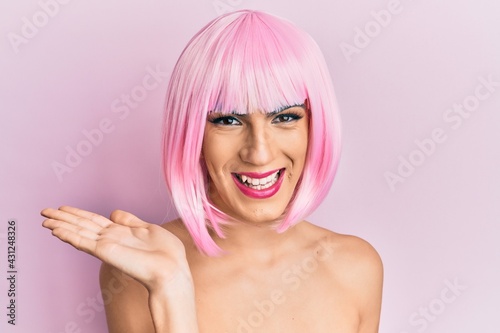 Young man wearing woman make up wearing pink wig smiling cheerful presenting and pointing with palm of hand looking at the camera.