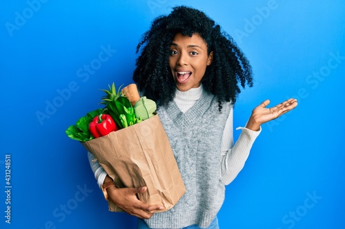 African american woman with afro hair holding paper bag with groceries celebrating achievement with happy smile and winner expression with raised hand photo