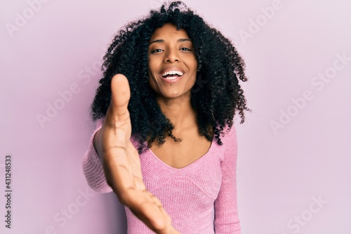 African american woman with afro hair wearing casual pink shirt smiling friendly offering handshake as greeting and welcoming. successful business.