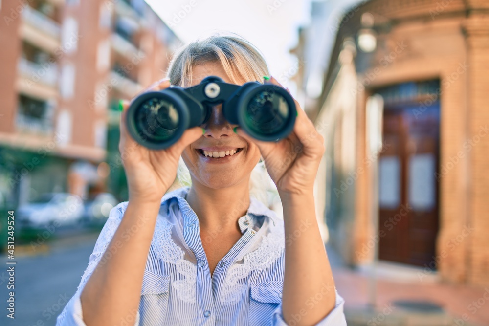 Young blonde woman smiling happy looking for new opportunity using binoculars at the city.
