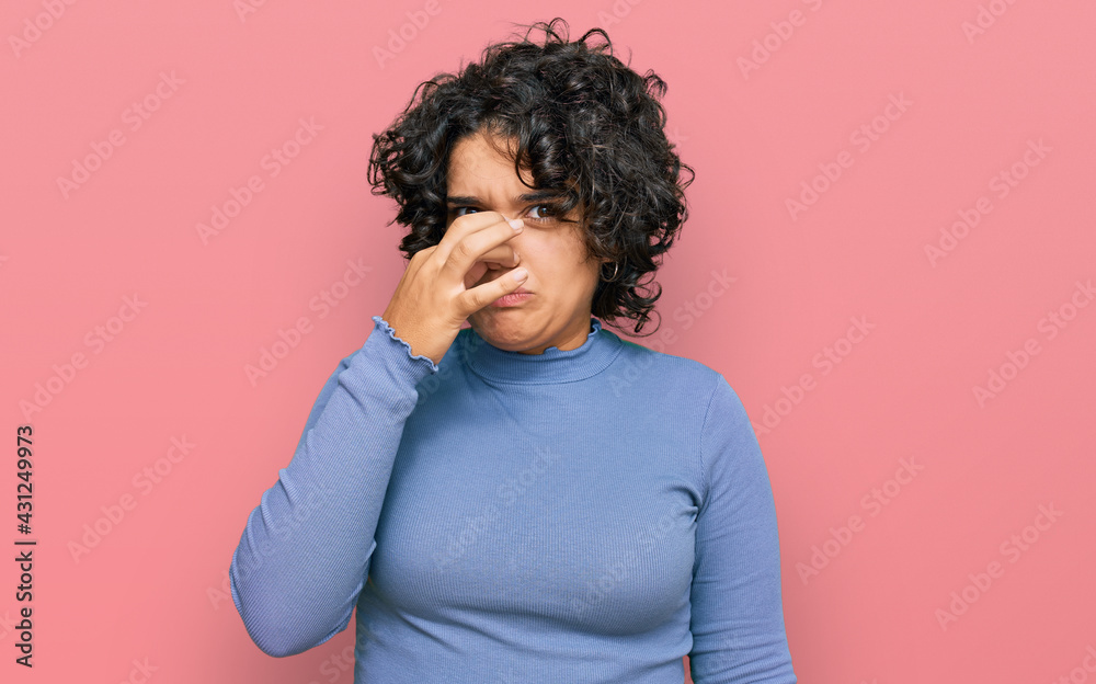 Young hispanic woman with curly hair wearing casual clothes smelling something stinky and disgusting, intolerable smell, holding breath with fingers on nose. bad smell Young hispanic woman with curly hair wearing casual clothes smelling something stinky and disgusting, intolerable smell, holding breath with fingers on nose. bad smell