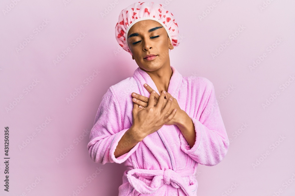 Hispanic man wearing make up wearing shower towel cap and bathrobe smiling with hands on chest with closed eyes and grateful gesture on face. health concept. Hispanic man wearing make up wearing shower towel cap and bathrobe smiling with hands on chest with closed eyes and grateful gesture on face. health concept.