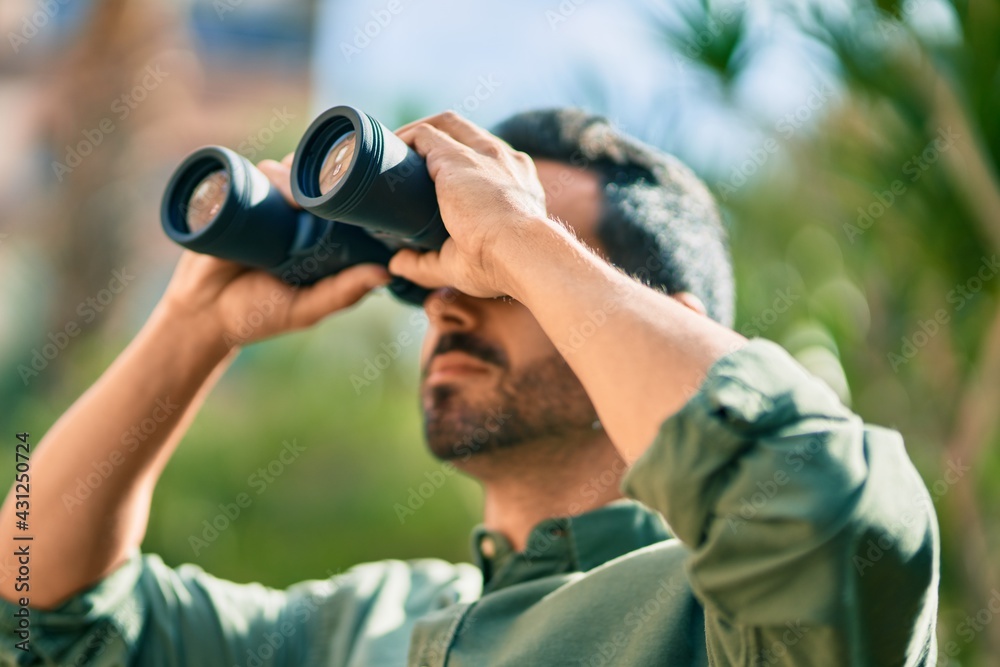 Young hispanic man smiling happy looking for new opportunity using binoculars at the park. Young hispanic man smiling happy looking for new opportunity using binoculars at the park.