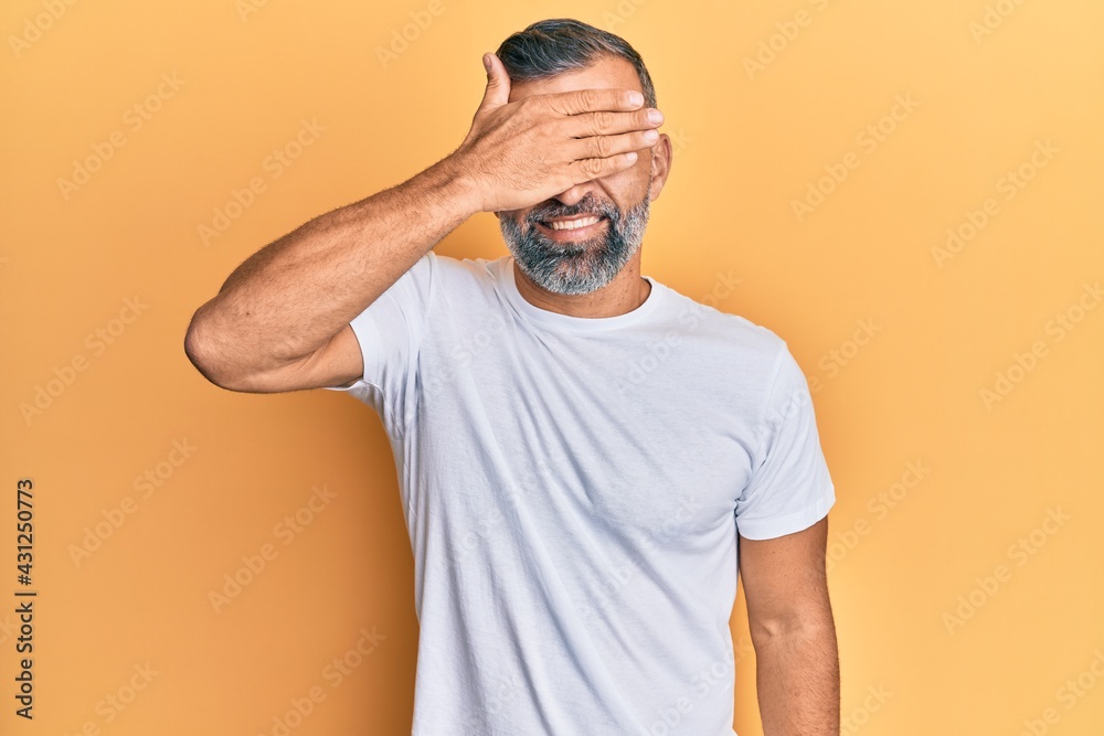 Middle age handsome man wearing casual white tshirt smiling and laughing with hand on face covering eyes for surprise. blind concept. Middle age handsome man wearing casual white tshirt smiling and laughing with hand on face covering eyes for surprise. blind concept.