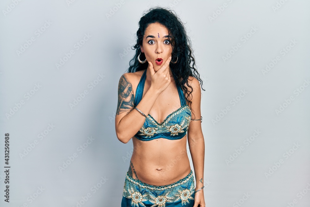 Young woman wearing bindi and traditional belly dance clothes looking fascinated with disbelief, surprise and amazed expression with hands on chin