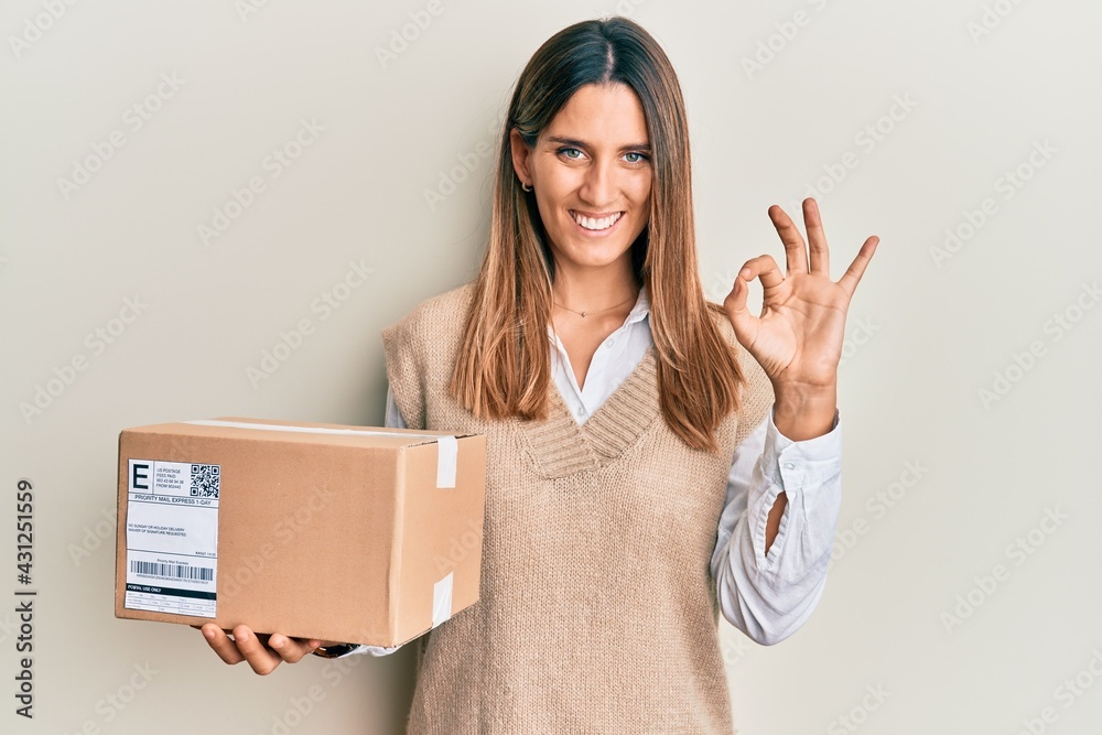 Brunette young woman holding delivery package doing ok sign with fingers, smiling friendly gesturing excellent symbol Brunette young woman holding delivery package doing ok sign with fingers, smiling friendly gesturing excellent symbol