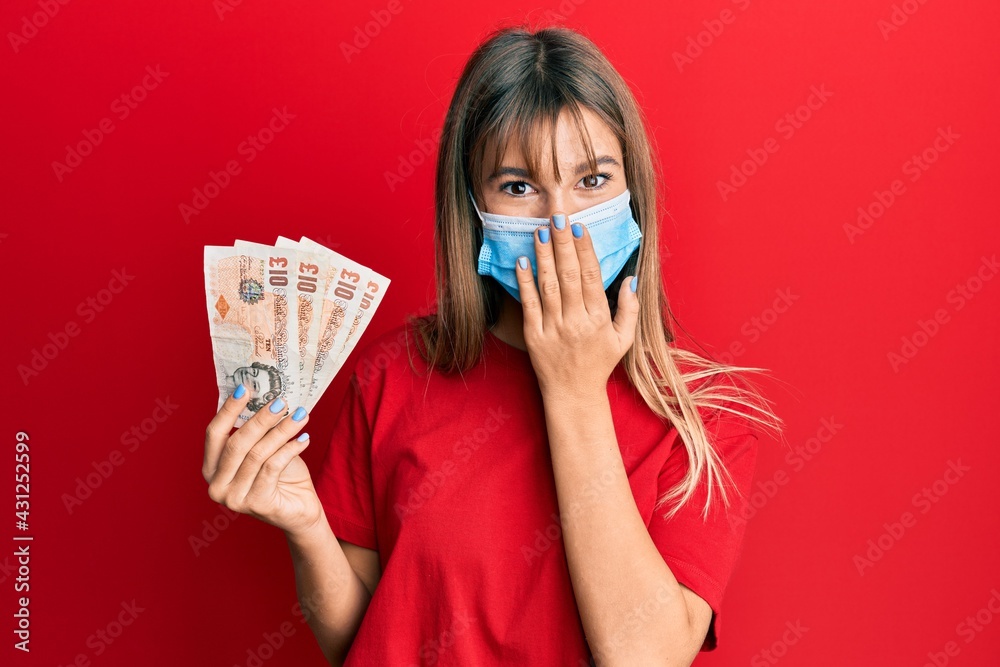 Teenager caucasian girl wearing medical mask holding 10 united kingdom pounds banknotes covering mouth with hand, shocked and afraid for mistake. surprised expression Teenager caucasian girl wearing medical mask holding 10 united kingdom pounds banknotes covering mouth with hand, shocked and afraid for mistake. surprised expression
