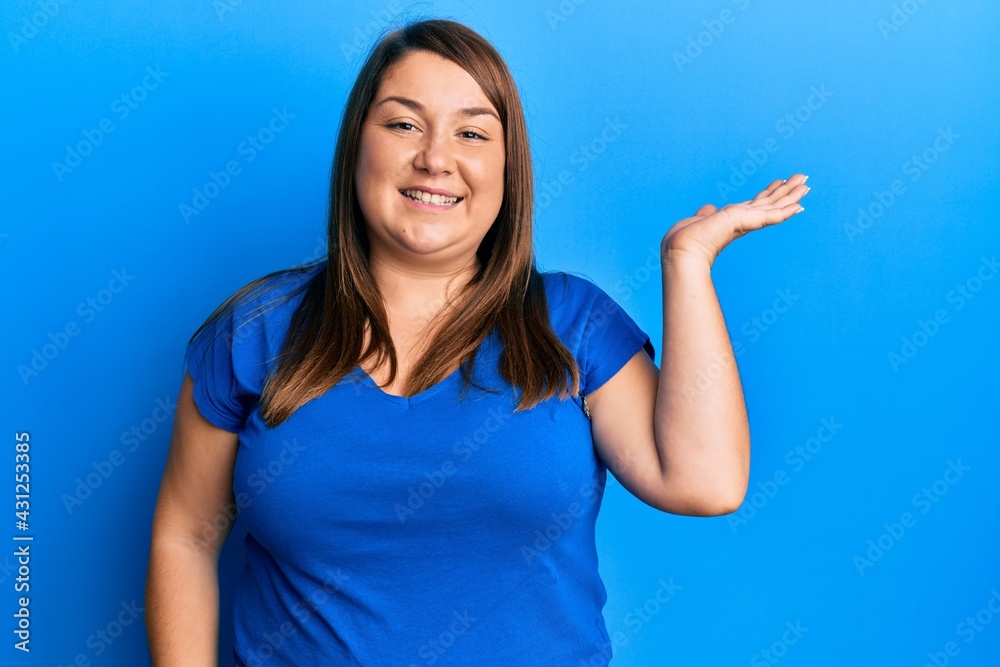 Fototapeta premium Beautiful brunette plus size woman wearing casual blue t shirt smiling cheerful presenting and pointing with palm of hand looking at the camera.