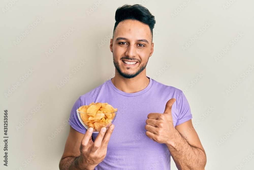 Young arab man holding potato chip smiling happy and positive, thumb up doing excellent and approval sign Young arab man holding potato chip smiling happy and positive, thumb up doing excellent and approval sign