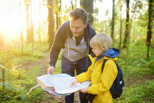Schoolchild and his mature father hiking together and exploring nature. Little boy with dad looking map during orienteering in forest. Adventure, scouting and hiking tourism for kids.
