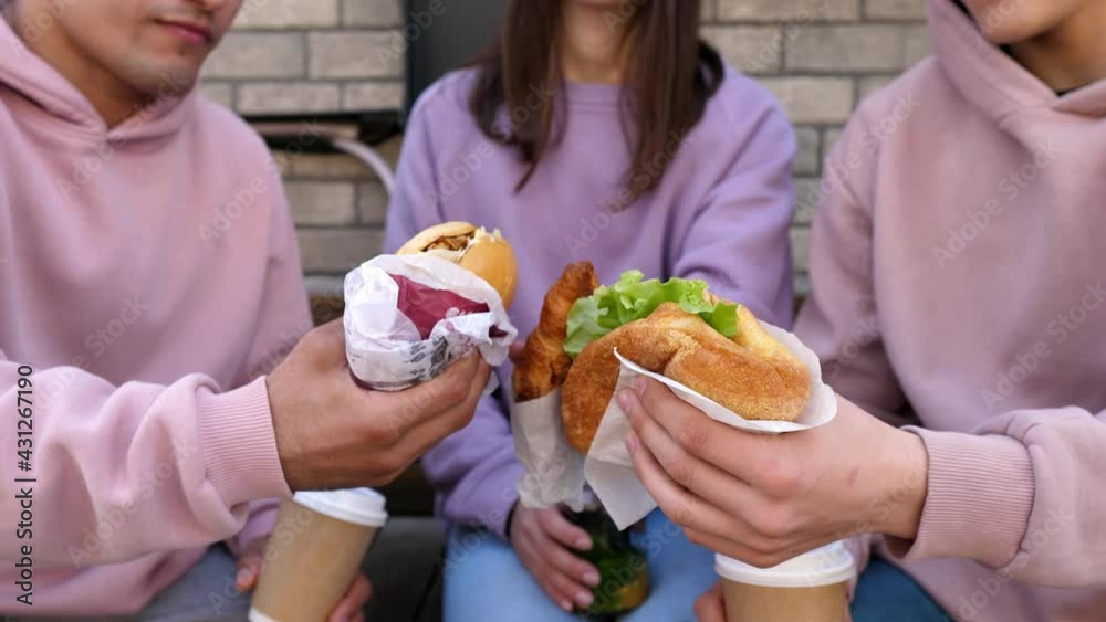 Group of young friends eating takeaway street food while sitting on a bench and chewing on vegan hamburgers. Three happy friends eating healthy food in the park