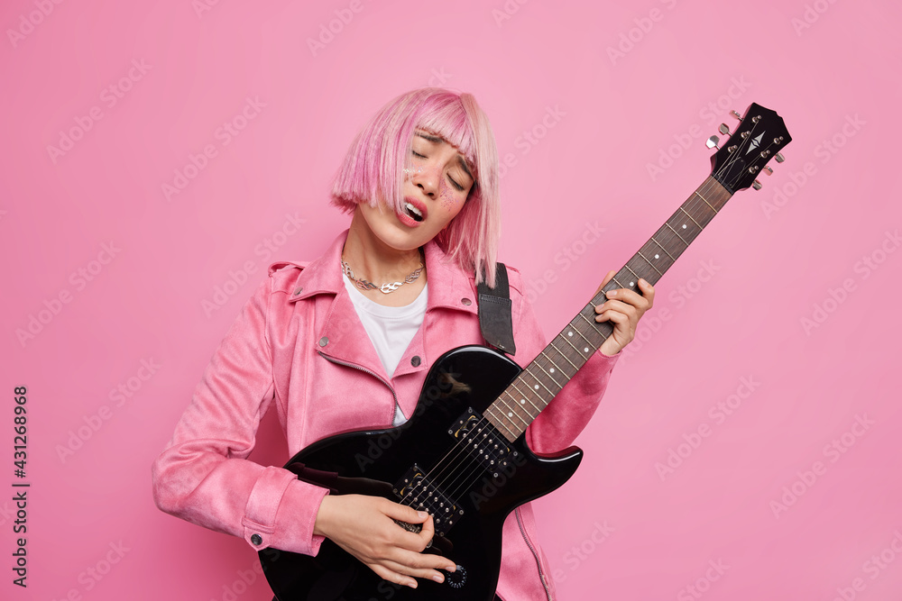 Carefree female musician tilts head sings favorite song plays electric guitar performs on stage being popular rock star jas trendy pink hair poses indoor. Music lifestyle show business concept