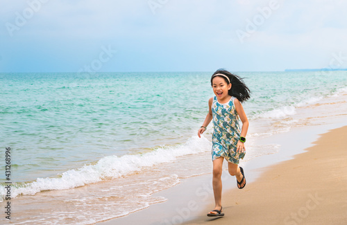 Happy little girl is running along the beautiful beach, motion blur, smiling face, wearing cute dress, black long hair, 7 years old, turquoise color ocean and blue sky. Space for copy and design.