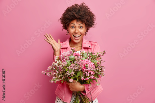 Portrait of good looking African American woman raises palm reacts on unexpected congratulations has date with boyfriend holds big bunch of flowers isolated over pink background. Holiday concept