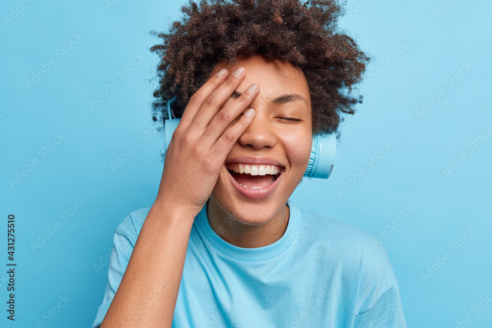 Positive currly haired woman makes face palm smiles happily has carefree expression listens audio track via headphones dressed in casual t shirt isolated over blue background. Emotions lifestyle Positive currly haired woman makes face palm smiles happily has carefree expression listens audio track via headphones dressed in casual t shirt isolated over blue background. Emotions lifestyle