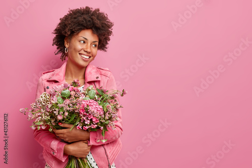 Studio shot of glad African American woman holds big bouquet of flowers celebrates spring holiday smiles gladfully looks away poses against pink background with copy space for your promotion