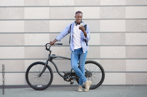 Cheerful African American man with bicycle standing near brick wall and choosing music playlist on smartphone