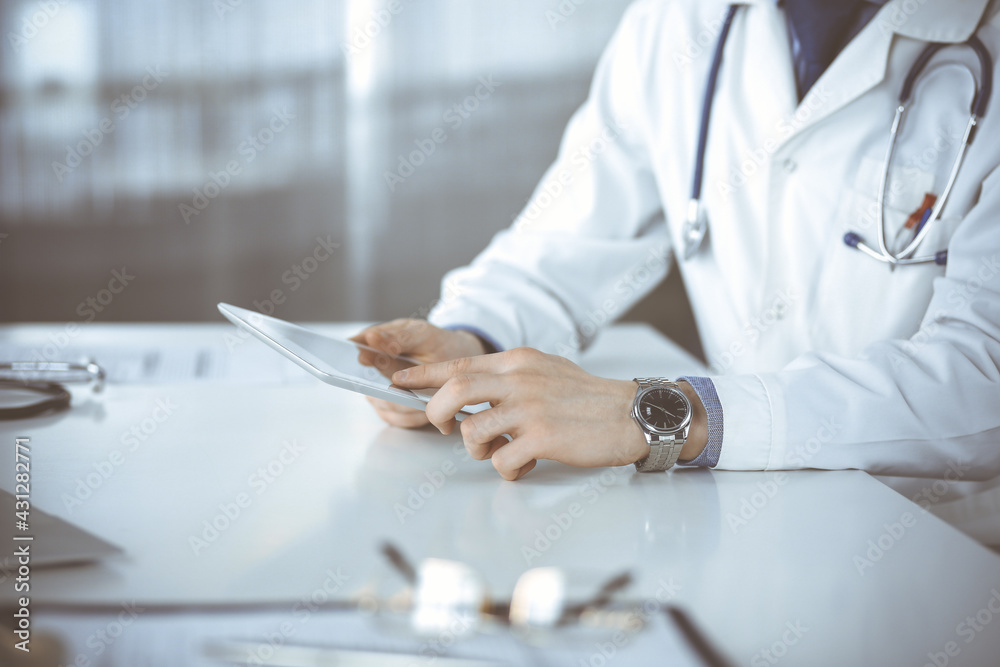 Unknown male doctor sitting and working with tablet computer in clinic at his working place, close-up. Young physician at work. Perfect medical service, medicine concept Unknown male doctor sitting and working with tablet computer in clinic at his working place, close-up. Young physician at work. Perfect medical service, medicine concept
