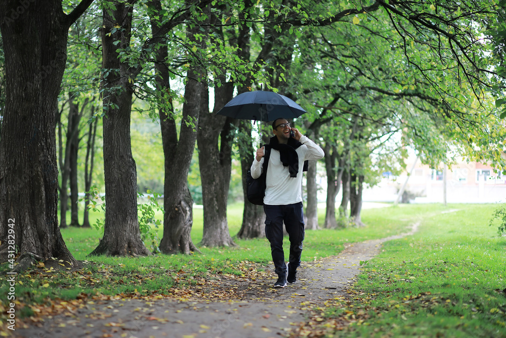 A young man in glasses walks in the park with an umbrella during the rain. A young man in glasses walks in the park with an umbrella during the rain.