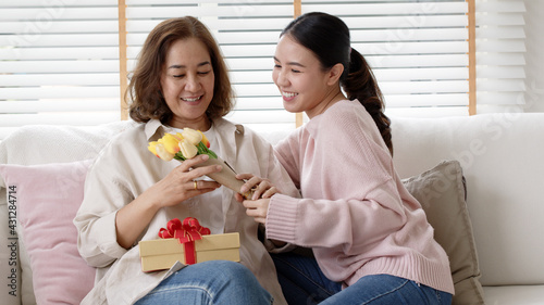 Attractive beautiful asian middle age mum sit with grown up daughter give gift box and flower in family moment celebrate mother day. Overjoy bonding cheerful kid embrace relationship with retired mom.