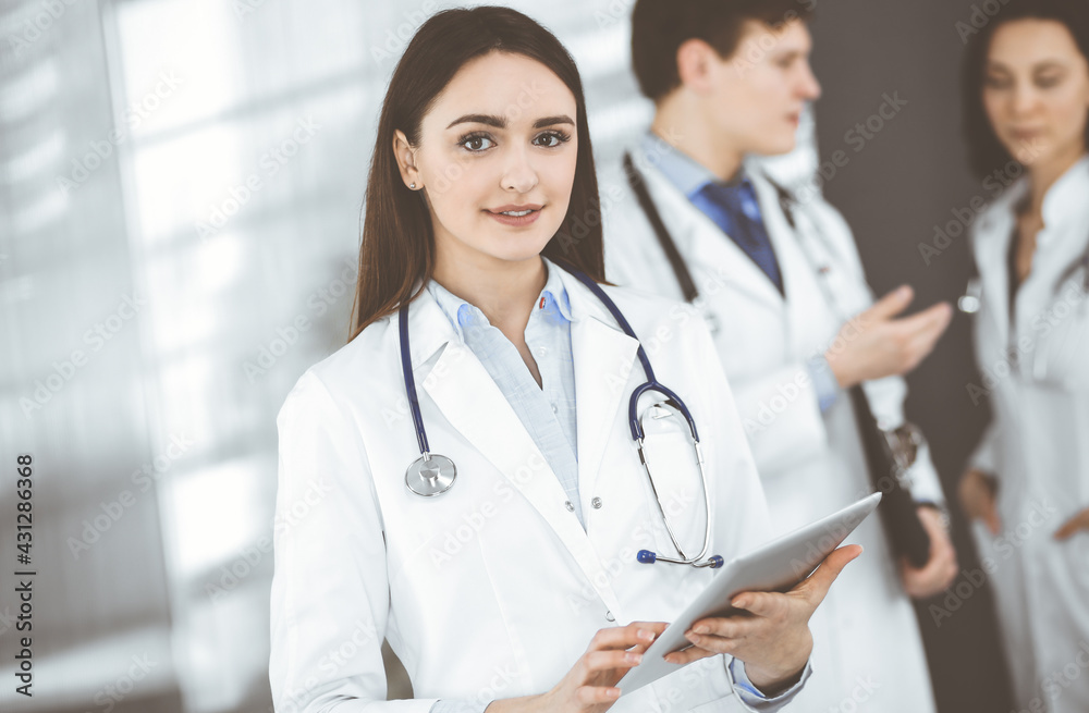 Smiling intelligent woman-doctor is holding a tablet computer in her hands, while she is standing together with her colleagues in a clinic. Physicians at work. Perfect medical service in a hospital Smiling intelligent woman-doctor is holding a tablet computer in her hands, while she is standing together with her colleagues in a clinic. Physicians at work. Perfect medical service in a hospital
