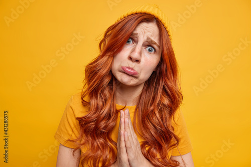 Upset young redhead woman purses lips keeps palms in pray begs you about something with pitiful expression looks hopeful dressed in casual yellow clothes has needy hopeless face stands indoor