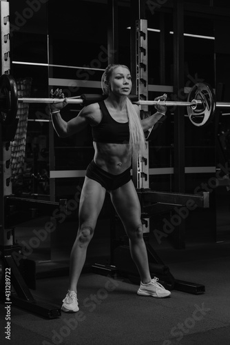 Fototapeta Naklejka Na Ścianę i Meble -  A sporty woman with blonde hair is squatting with a barbell near the squat rack in a gym. A girl is doing a leg workout.