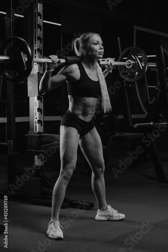 A sporty woman with blonde hair is started squatting with a barbell near the squat rack in a gym. A girl is doing a leg workout.