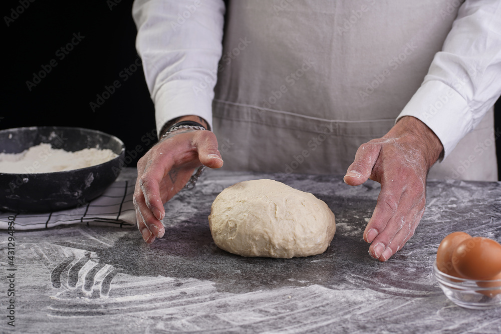A male chef baker in a white shirt and grey apron kneading dough on a dark kitchen table