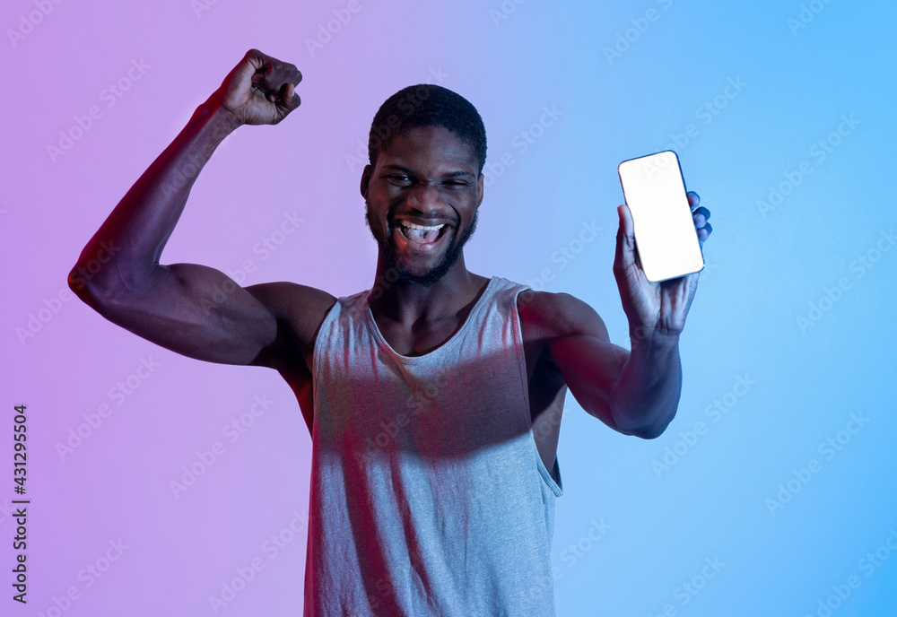 Fototapeta premium Excited black athlete showing cellphone with blank screen, making YES gesture in neon light, mockup for design