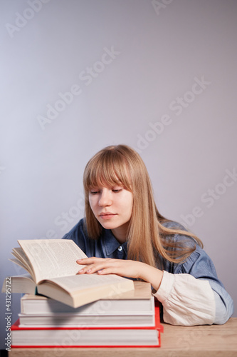 Young beautiful caucasian pupil female with books in class with copy space. Attractive caucasian happy blonde student girl sitting at the table reading book. High quality vertical photo