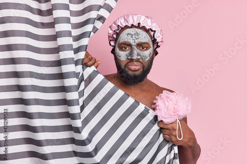 Dissatisfied Afro American man with fresh dark skin takes care of body and skin takes shower regularly hold bath sponge applies clay mask on face poses behind striped curtain. Persoonal hygiene photo