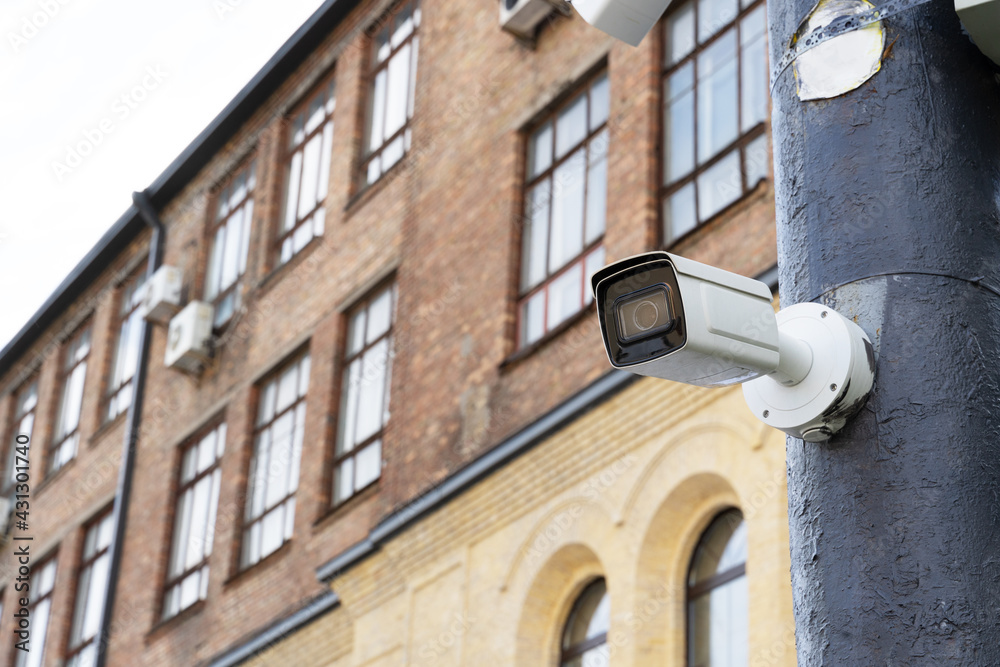 White surveillance camera on a gray pole on a blurred brick house background. Security observation, protection against offenses White surveillance camera on a gray pole on a blurred brick house background. Security observation, protection against offenses