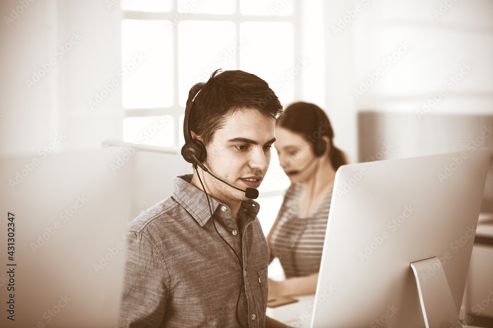 Casual dressed young man using headset and computer while talking with customers online. Group of operators at work. Call center Casual dressed young man using headset and computer while talking with customers online. Group of operators at work. Call center