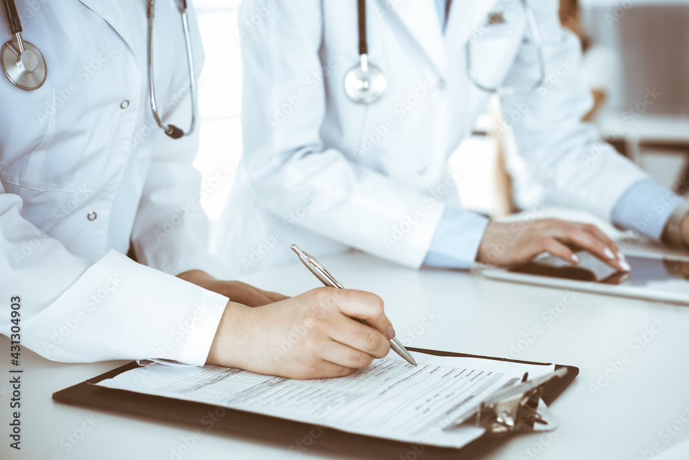 Unknown woman-doctors at work with patient at the background. Female physicians filling up medical documents or prescription while standing in hospital reception desk, close-up. Health care concept Unknown woman-doctors at work with patient at the background. Female physicians filling up medical documents or prescription while standing in hospital reception desk, close-up. Health care concept