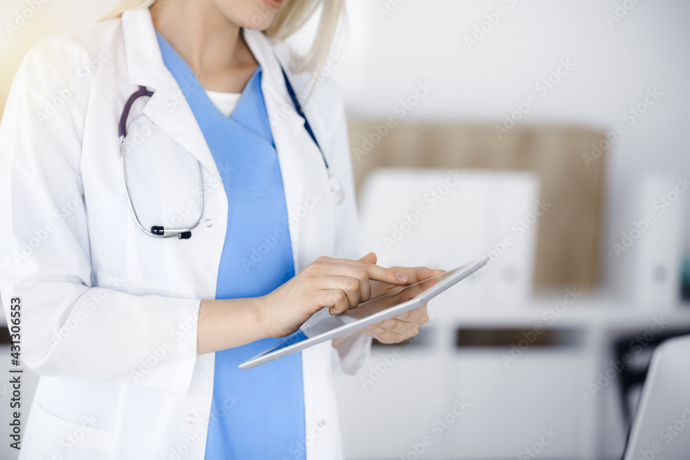 Unknown woman-doctor standing in sunny clinic and using tablet pc, close-up. Data and best service in medicine Unknown woman-doctor standing in sunny clinic and using tablet pc, close-up. Data and best service in medicine
