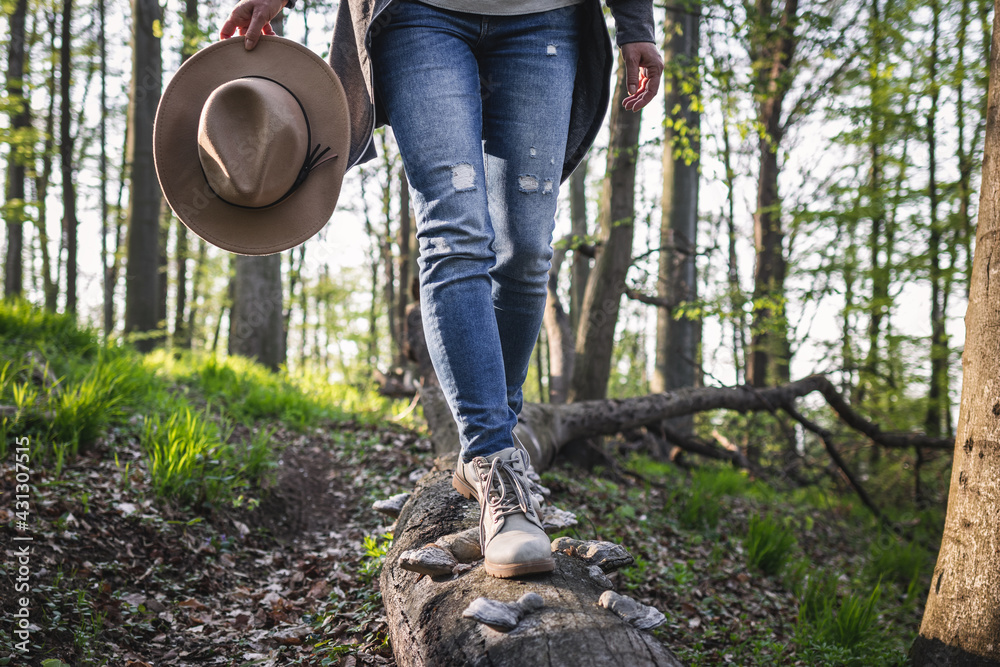 Hiking in forest. Woman wearing jeans, holding hat and walking on tree trunk. Active lifestyle in nature - obrazy, fototapety, plakaty 