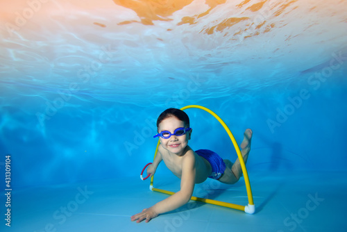A beautiful little boy swims and poses underwater at the bottom of a children's pool. He looks at the camera and smiles as he swims through the hula hoop. Portrait. Concept. Horizontal view