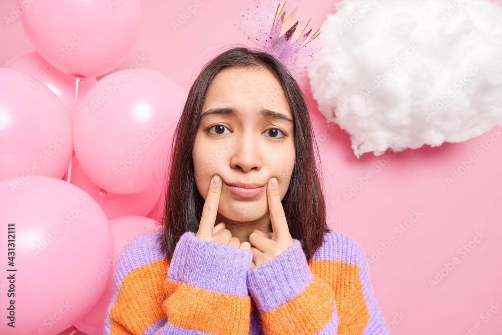 Sad dejected Asian woman has dark hair makes smile with fingers being in bad mood on birthday party wears princess crown on head warm sweater poses against decorated background near bunch of balloons Sad dejected Asian woman has dark hair makes smile with fingers being in bad mood on birthday party wears princess crown on head warm sweater poses against decorated background near bunch of balloons