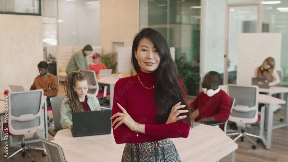 Portrait slowmo of confident young Asian businesswoman posing with her arms crossed in open-plan office. She is smiling and looking at camera while businesspeople working in background Portrait slowmo of confident young Asian businesswoman posing with her arms crossed in open-plan office. She is smiling and looking at camera while businesspeople working in background
