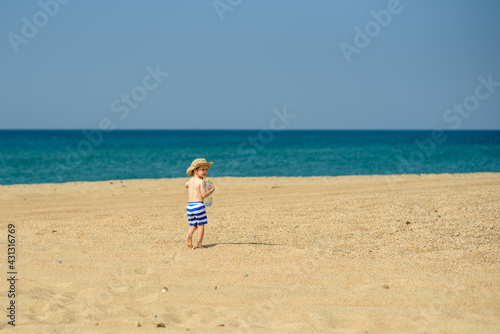 A small boy alone plays on the seashore, wearing a hat and striped shorts. Heat