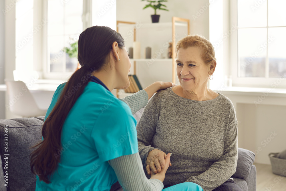 Friendly doctor supporting senior woman. Happy young nurse or clinician and her older adult patient sitting on sofa, looking at each other, holding hands, talking and smiling. Health and care concept Friendly doctor supporting senior woman. Happy young nurse or clinician and her older adult patient sitting on sofa, looking at each other, holding hands, talking and smiling. Health and care concept