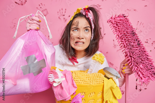 Cleaning concept. Puzzled Asian girl helps mother to do work about house holds garbage bag dirty mop has frustrated tired face expression looks upwards stands near basket with detergents and laundry