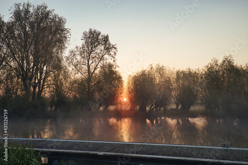 Horizontal view on small river Linge during sunrise in spring with a pier, trees and willows on the horizon, morning haze and reflections on the water. Betuwe in Netherlands with copy space.