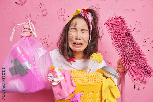 Frustrated displeased housemaid cries unhappily being tired of house cleaning holds garbage bag and mop collects all dirty laundry isolated over pink background. Daily home routines concept.