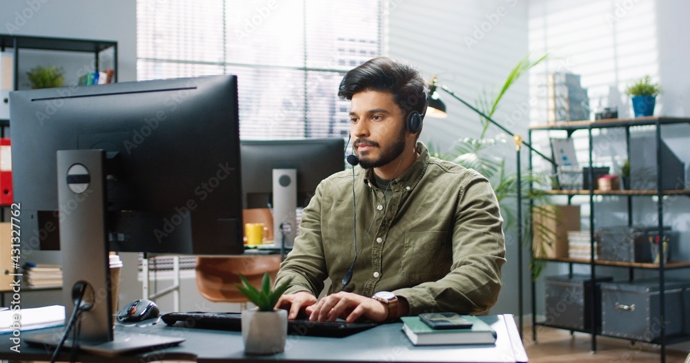 Portrait of joyful handsome young Hindu male worker in headset sitting at table in modern cabinet and texting on laptop browsing online at office looking at camera and smiling. Business concept Portrait of joyful handsome young Hindu male worker in headset sitting at table in modern cabinet and texting on laptop browsing online at office looking at camera and smiling. Business concept