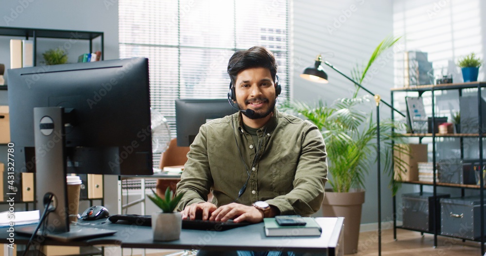 Portrait of joyful handsome young Hindu male worker in headset sitting at table in modern cabinet and texting on laptop browsing online at office looking at camera and smiling. Business concept Portrait of joyful handsome young Hindu male worker in headset sitting at table in modern cabinet and texting on laptop browsing online at office looking at camera and smiling. Business concept