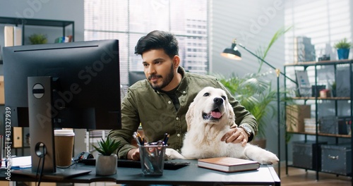 Photography Portrait of joyful Hindu handsome man sitting in cabinet at work with his cute dog pet in good mood typing on computer browsing online